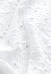 Witte stof met ingewikkeld borduurwerk met cirkelvormige uitsparingen en verhoogde patronen, die een textuurrijk en delicaat ontwerp tonen.