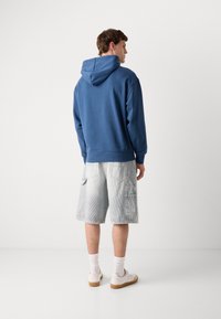 Felpa blu con cappuccio e tasca a marsupio, abbinata a pantaloni cargo grigi chiari con due tasche laterali, indossata con scarpe da ginnastica bianche.