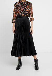 Svart plisserad midikjol som matchas med en genomskinlig svart blus med blommor i rosa och orange. Modellen bär svarta ankelboots.