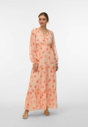 Femme debout portant une longue robe pêche à imprimé floral orange, col en V, taille froncée, manches longues et jupe à volants, chaussant des sandales.