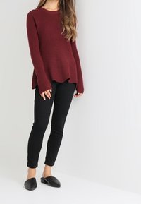 Donna in piedi che indossa un maglione bordeaux a coste, pantaloni neri attillati e scarpe slip-on nere a punta, su uno sfondo semplice.