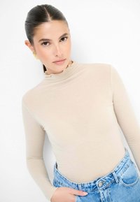 Top beige a maniche lunghe con colletto alto a volant, abbinato a jeans di denim azzurri a vita alta. Tessuto liscio e design aderente.