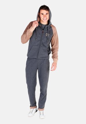 Jeune homme souriant portant un sweat à capuche gris et marron à fermeture éclair, pantalon gris assorti et baskets blanches, debout devant un fond uni.