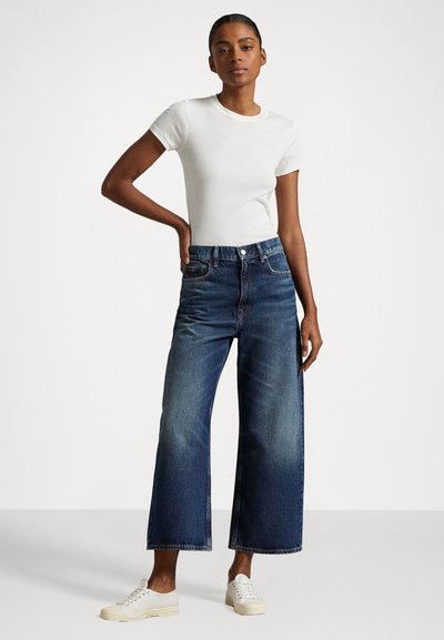 Λευκό φανελάκι, τζιν σε μέτρια απόχρωση με φαρδιά πόδια, κοντό μήκος, μαλακή υφή, σε συνδυασμό με λευκά αθλητικά παπούτσια. Καθαρή, καθημερινή εμφάνιση.