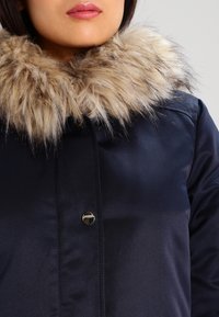 Žena v námořnickém modrém zimním kabátu s tlustým béžovým a hnědým kožešinovým límcem, viditelná pouze od rtů a spodní části obličeje.