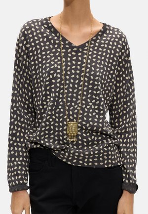 Kobieta w czarno-białym swetrze w groszki z dekoltem w serek i długimi rękawami, czarnych spodniach oraz długim złotym łańcuszku z prostokątnym wisiorkiem.