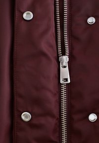 Burgunderfarbene Jacke mit einem silbernen Metallreißverschluss, Druckknöpfen und einer glatten Stoffstruktur mit leichtem Glanz.