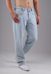 Mann trägt hellblaue Jeans und weiße Sneakers, steht mit einem Bein leicht gebeugt und einer Hand in der Tasche.