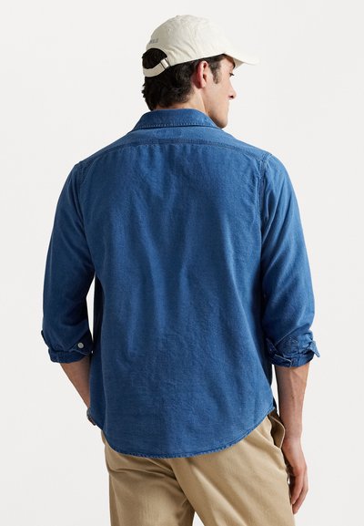 Blue denim shirt with rolled sleeves, buttoned cuffs, and a curved hem. The back features a plain texture and a classic collar design.