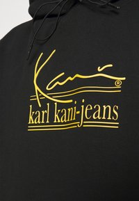 Mustapaita, jossa on kirkas keltainen logo, jossa on tyylitelty teksti "karl kani-jeans" ja koristeelliset viivat tekstin ylä- ja alapuolella.