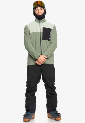 Chaqueta de forro polar en verde con detalles en negro, cierre completo y un bolsillo en el pecho; combinada con pantalones de nieve negros y botas de esquí.