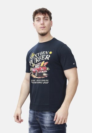 Giovane uomo che indossa una T-shirt nera con una grafica di un camion rosso e tavole da surf e la scritta "Next Gen Surfer", abbinata a jeans blu slavati.