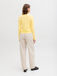 Vrouw met schouderlang bruin haar, gekleed in een gele trui met lange mouwen, lichtbeige broek met wijde pijpen en zwarte schoenen, staand met haar rug naar de kijker gekeerd.