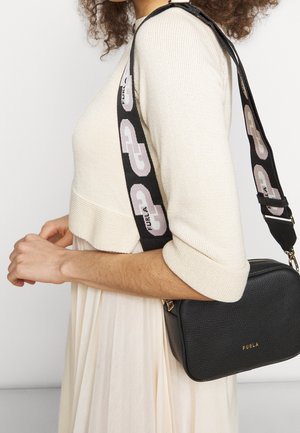 Black textured leather crossbody bag with gold hardware and a patterned strap featuring “Furla” logo, worn over a light dress with a cropped sweater.