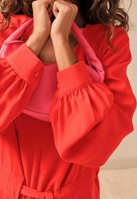 Blouse rouge à manches bouffantes, poignets pliés et ceinture. Tenant un sac à main rose à la texture lisse et à la forme structurée.