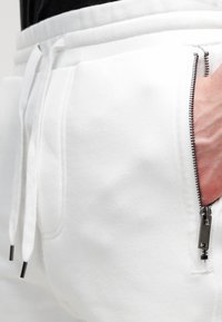 Bílé tepláky vyrobené z měkké tkaniny, s kapsou na zip a kovovým kováním a nastavitelnou šňůrkou v pase.