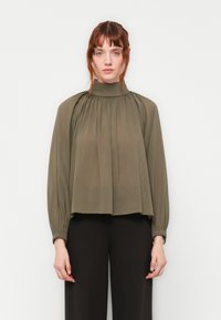 Kvinne med rødt hår iført en olivenfarget bluse med høy hals og gathered-ermer, samt svarte vide bukser, som står foran en ensfarget bakgrunn.