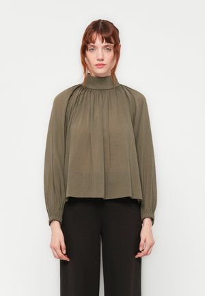 Kvinne med rødt hår iført en olivenfarget bluse med høy hals og gathered-ermer, samt svarte vide bukser, som står foran en ensfarget bakgrunn.