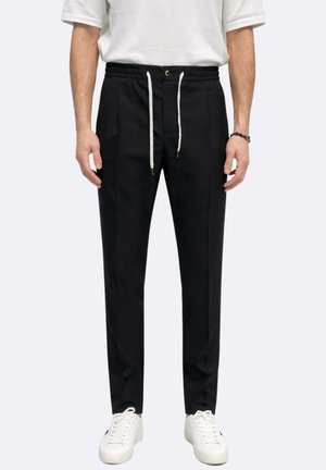 Uomo che indossa pantaloni neri su misura con lacci bianchi, sneakers bianche e una maglietta bianca a maniche corte, in piedi contro uno sfondo uniforme.