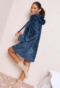 Niebieski, oversize'owy szlafrok z kapturem wykonany z miękkiej tkaniny, ozdobiony złotym haftem przedstawiającym słońce i księżyc, o luźnym kroju. Założone beżowe skarpetki.