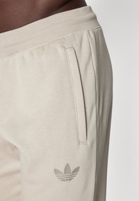Helle beige Jogginghosen aus weichem Stoff, ausgestattet mit seitlichen Taschen und einem dezenten grauen Adidas-Logo am linken Bein. Mit elastischem Bund.