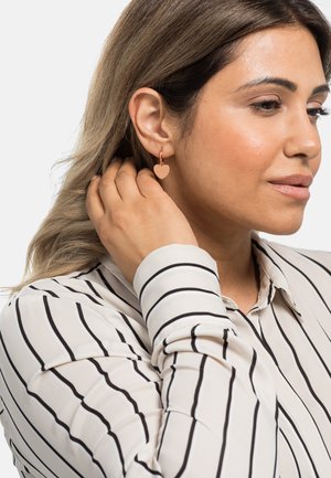Ohrringe in Herzform aus Roségold mit gerippter Textur, die an einem Hoops befestigt sind. Die Trägerin hat glattes, schulterlanges Haar und trägt ein gestreiftes Hemd.