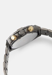 Reloj cronógrafo con caja de acero inoxidable, correa negra y acentos dorados en la corona y los pulsadores. Presenta un bisel texturizado y una parte trasera lisa.