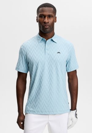 Mann trägt hellblaues, strukturiertes Kurzarm-Poloshirt und weißen Golfhandschuh an der rechten Hand vor einfarbigem Hintergrund.