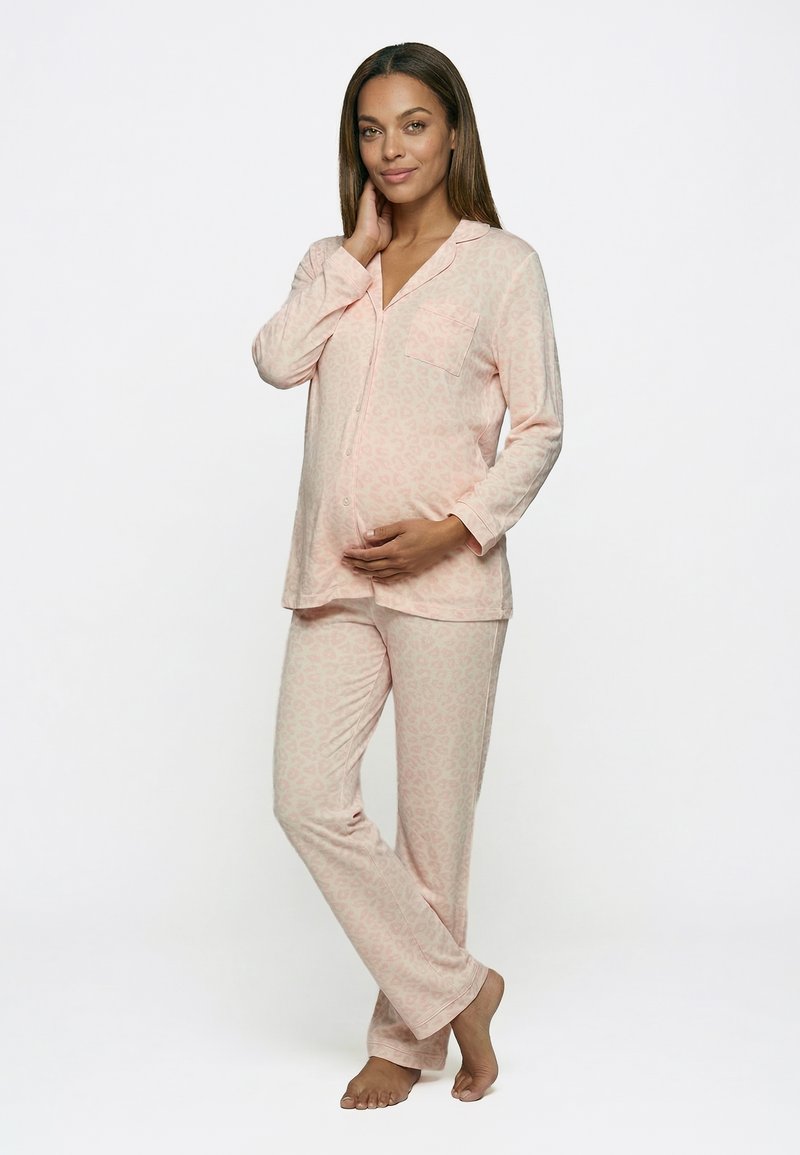 Femme enceinte en pyjama à manches longues rose clair à motifs, pieds nus, tenant doucement son ventre d'une main, debout sur fond blanc.