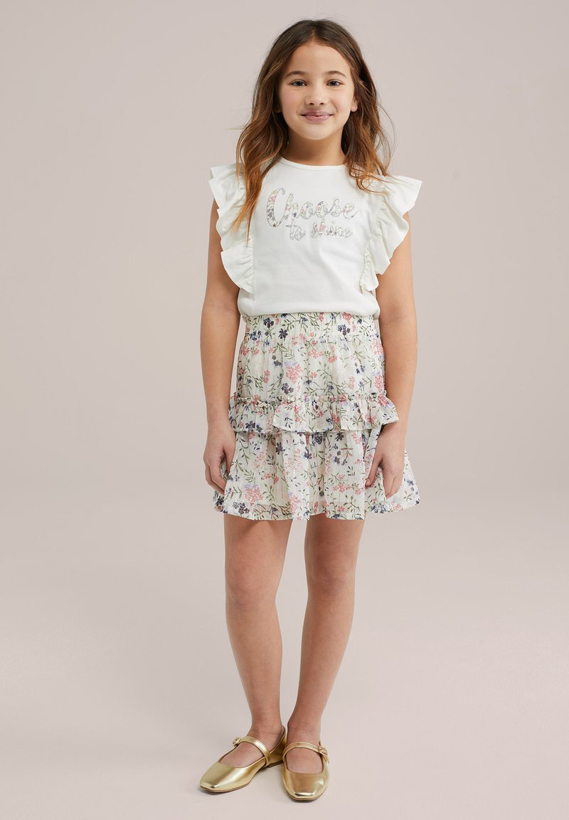 Balta trumpa rankovėmis palaidinė su raukiniais ir tekstu "Pasirink spindėti", derinama su gėlėta, sluoksniuota mini sijonu ir auksinėmis butų bateliais.