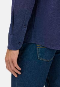 Mörkblå långärmad skjorta med strukturerad tyg, dubbelknapp på ärmsluten, i kombination med mörkblå jeans med gula sömmar på fickorna.