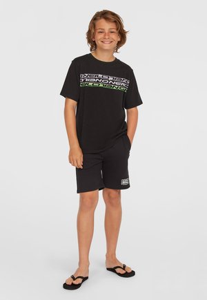 Sort t-shirt med grøn og hvid tekstgrafik, parret med sorte shorts. Outfitten inkluderer sorte flip-flops. Afslappet pasform og design.