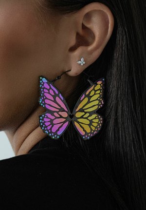 Großer, bunter Ohrring in Form eines Schmetterlings mit rosa, lila und gelben Flügeln, getragen am linken Ohr, darüber ein kleiner Blumenstecker.