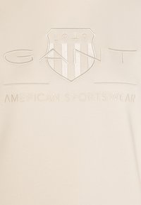 Felpa beige con logo in rilievo che presenta un design a scudo e il testo "GANT AMERICAN SPORTSWEAR". Texture liscia con un leggero lucido.