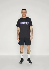 Crna pamučna majica s šarenim grafičkim printom na prsima, uparena s crnim kratkim hlačama, čarapa do gležnja i crnim sportskim cipelama.