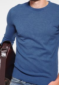 Pull bleu à col rond en tissu tricoté doux, coupe ajustée, porté avec un jean en denim, avec un sac bandoulière en cuir marron.
