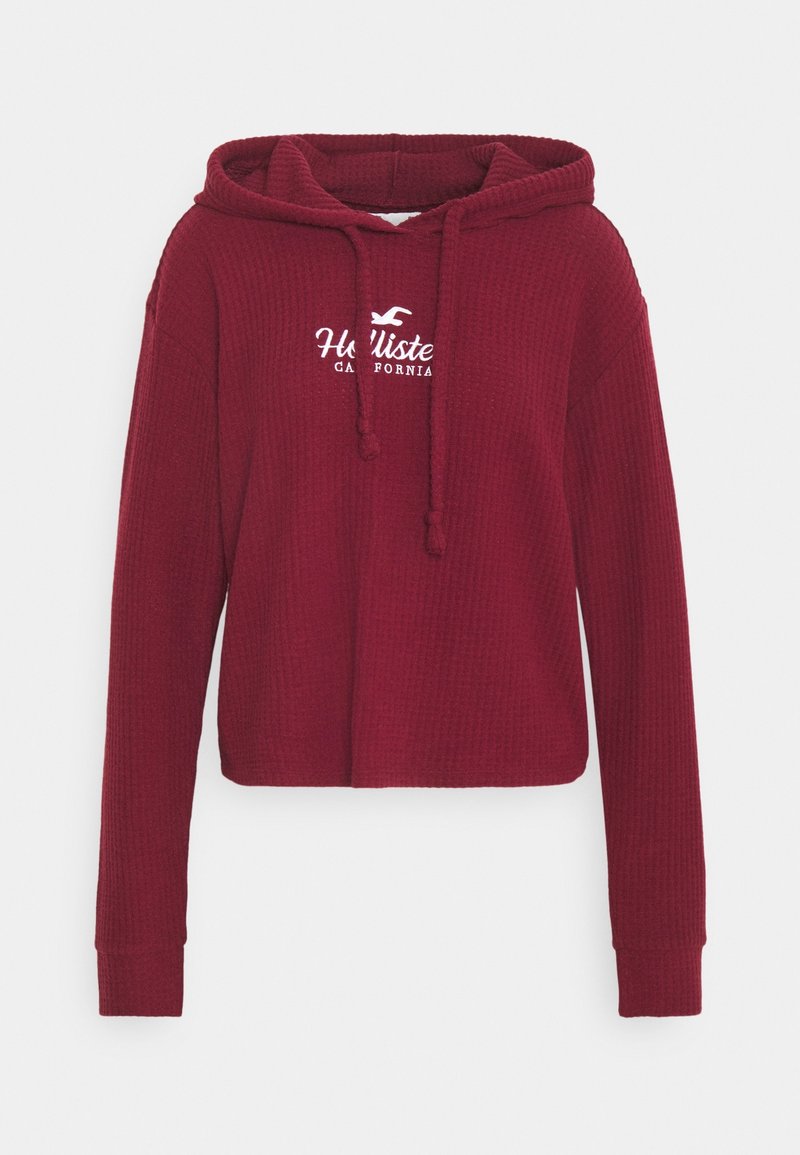 Burgundy lyhyttoppinen huppari, joka on valmistettu teksturoidusta kankaasta. Siinä on edessä kengurulompakko ja valkoinen brodeerattu logo rinnassa.
