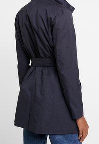 Trench coat navy con vita abbottonata, pieghe posteriori e colletto alto. Presenta tessuto liscio e leggero e maniche lunghe.