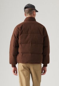 Ruskea, pörröinen fleecetakki, jossa on pystokaulus ja toppaustehokas malli. Siinä on resorit hihansuissa ja rento istuvuus. Yhdistetty khakivärisiin housuihin.