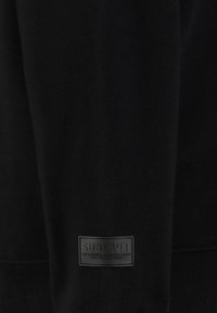 Schwarzer Sweatshirt aus weichem Stoff, mit einem strukturierten Logo-Patch an der unteren Vorderseite, auf dem der Markenname "SUBLEVEL" in erhabenen Buchstaben zu sehen ist.