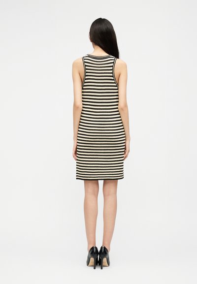 Ženska z dolgimi temnimi lasmi, oblečena v brezrokavno črno-belo obleko s črtami do kolen in črne visoke pete, stoji obrnjena stran.