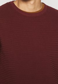 Bordeauxrode textuursweater met horizontale geribbelde details, ronde hals en een contrasterende bruine binnenkraag. Gemaakt van een katoenmix.