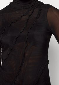 Svart genomskinlig topp med volangdetaljer längs framsidan och axeln. Har hög hals och långa ärmar, tillverkad av lätt, texturerat material.