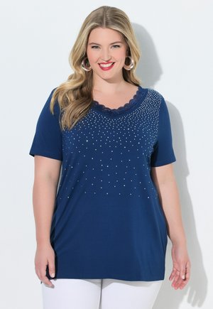 Smilende kvinde med langt blondt hår iført navyblå kortærmet bluse med rhinsten og hvide bukser, stående op ad en hvid væg.