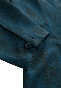 Tmavě tyrkysová látka s jemným květinovým vzorem, texturovaný povrch a detail knoflíku na manžetě. Hladký a elegantní design.