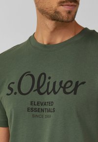 Zelené bavlnené tričko s okrúhlym výstrihom a čiernym potlačeným textom: "s.Oliver ELEVATED ESSENTIALS SINCE 1969."