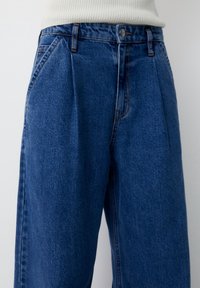 Denimbyxor har en djupblå färg med en strukturerad yta, hög midja med veck och ett slätt midjeband med en metalldekorativeknapp.