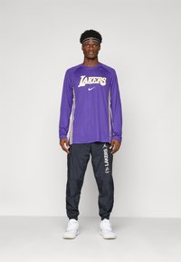 Mand iført en lilla langærmet Lakers-trøje, sorte Lakers-træningsbukser, hvide sneakers og et sort Nike-hovedbånd, stående mod en ensfarvet baggrund.
