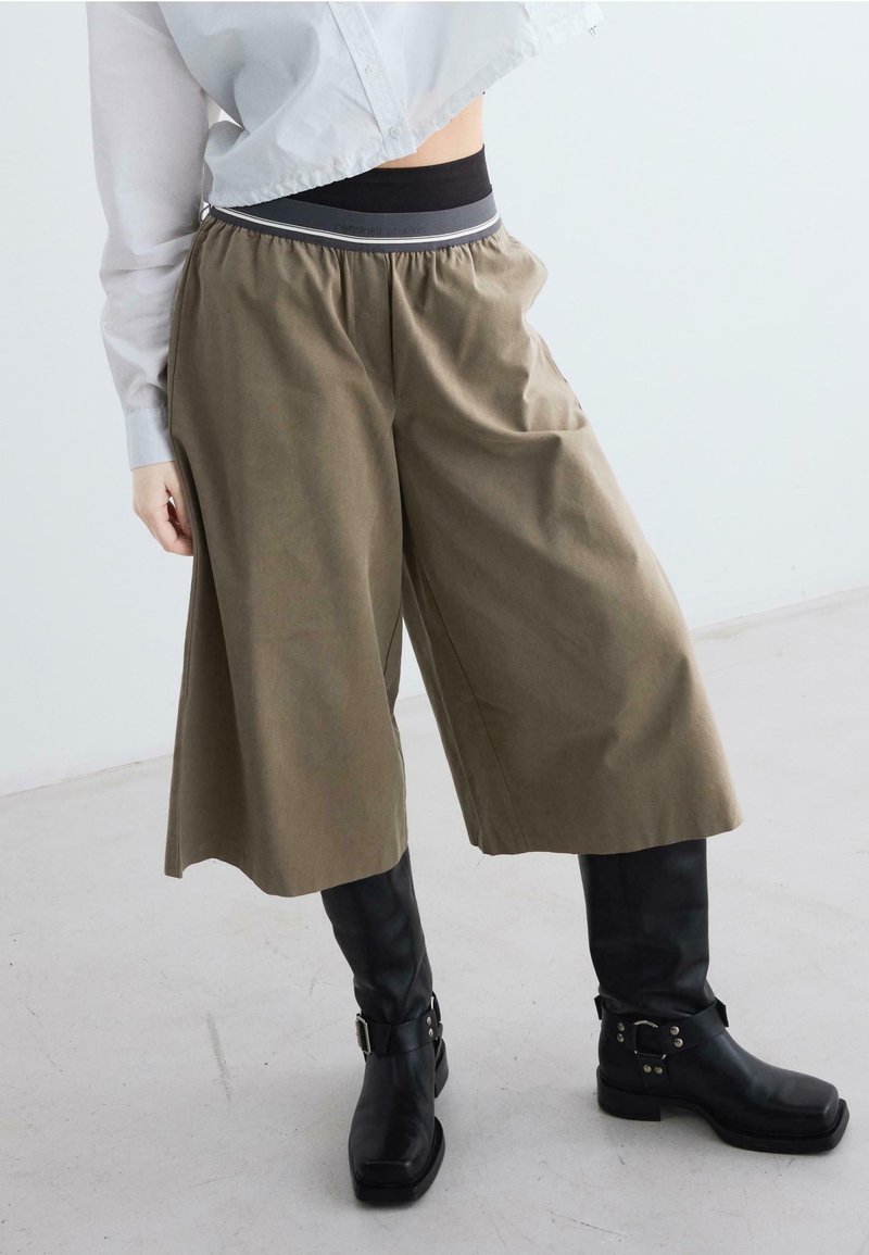 Person, die lockere olivgrüne Culottes, schwarze knöchelhohe Stiefel mit Schnallenriemen und ein kurzes hellgraues Shirt trägt, steht auf einem grauen Boden.