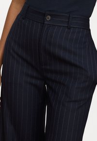 Pantaloni navy a righe sottili con vita aderente, dotati di chiusura con bottone anteriore e silhouette slanciata. Realizzati in tessuto morbido.